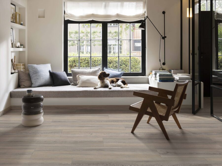 Woonkamer met pvc-vloer in warme houtlook, stalen deuren, knusse vouwgordijnen en hondje op de bank.