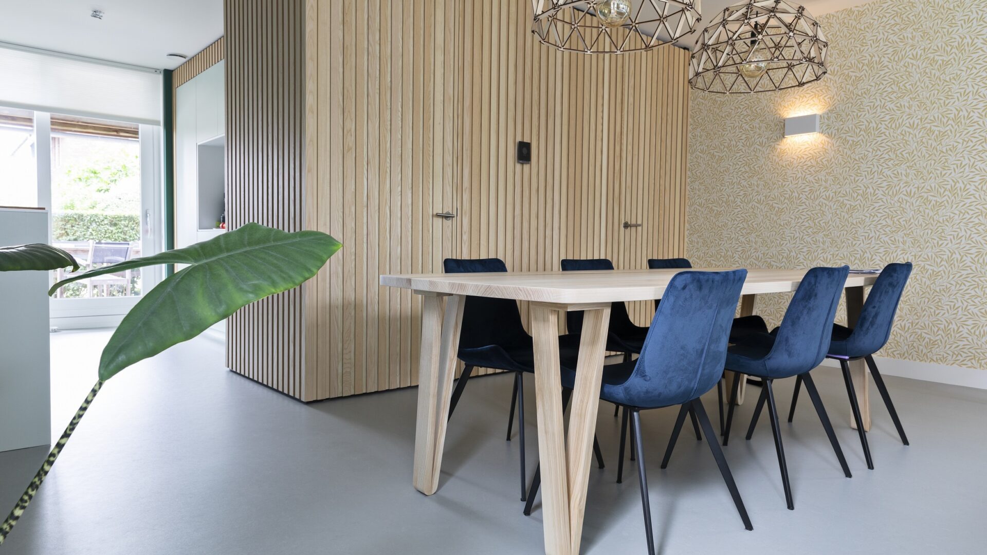 Eetkamer met grijze betonlook marmoleum vloer, houten tafel, stoelen met metalen poten en houten wandpanelen aan de muur