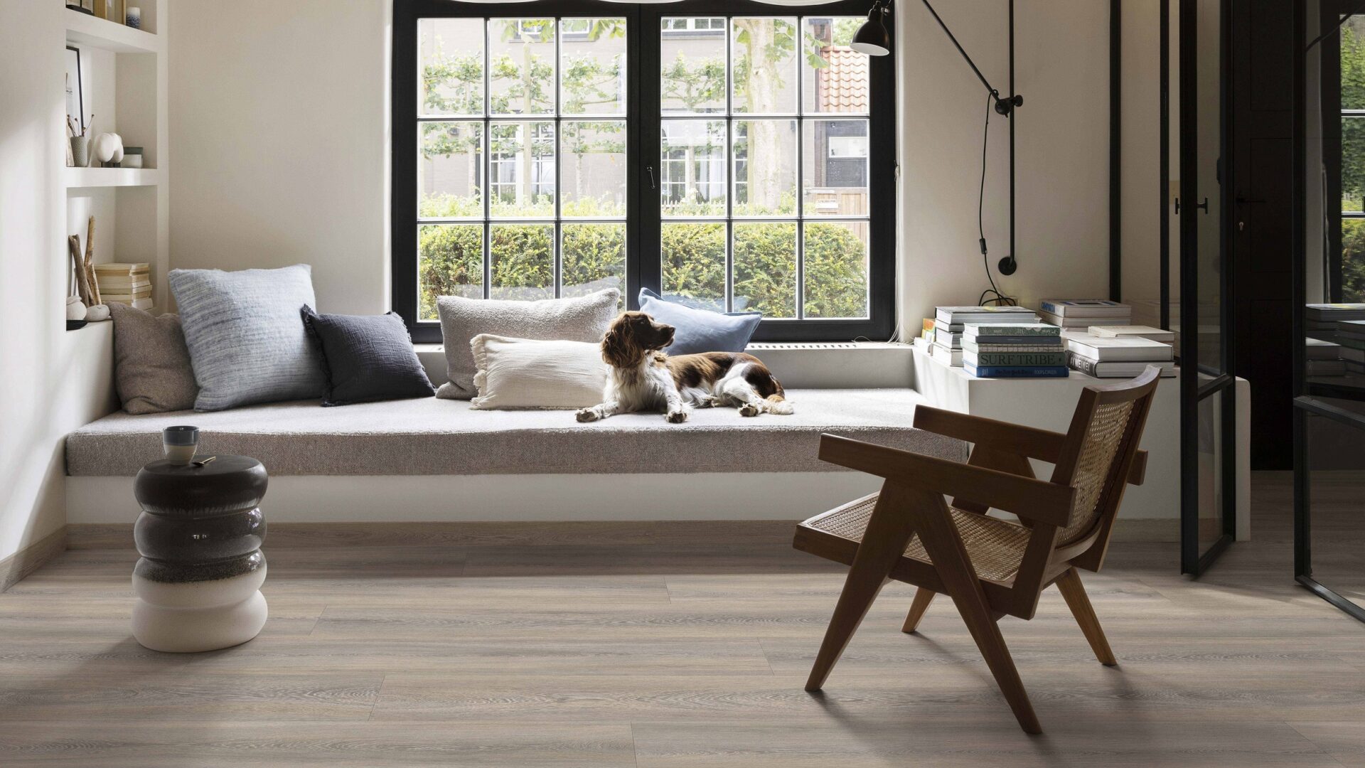 Woonkamer met pvc-vloer in warme houtlook, stalen deuren, knusse vouwgordijnen en hondje op de bank.