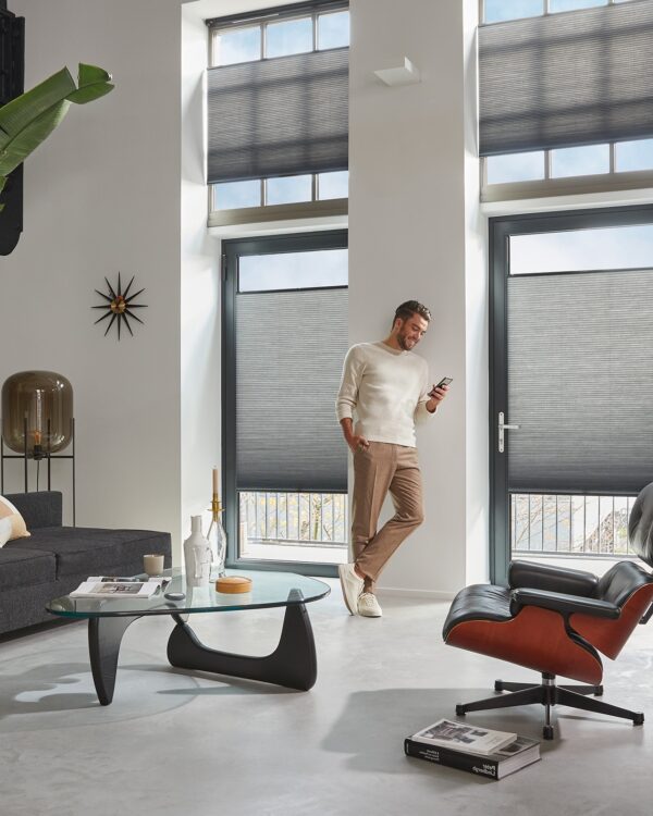 Grijze duette® shades van Luxaflex® in de woonkamer met man op telefoon