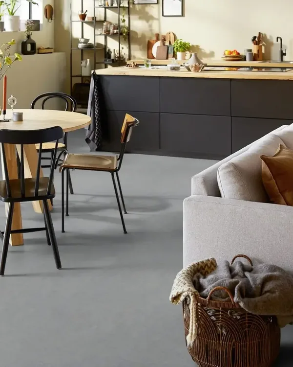Open woonkeuken met grijze marmoleumvloer, ronde houten eettafel, lichte bank en zwart keukeneiland