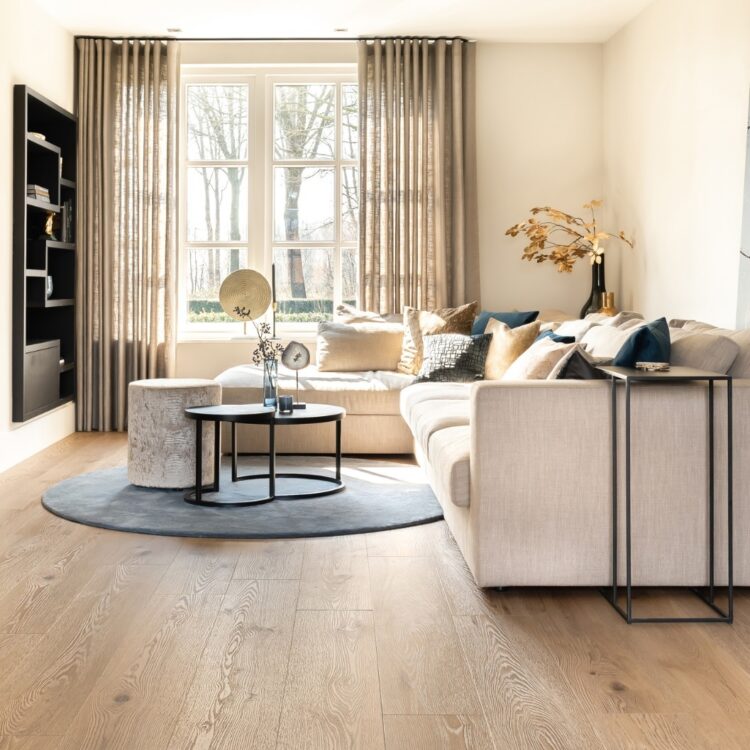 Woonkamer met witte hoekbank, stalen salontafel, transparante gordijnen en houten pvc-vloer met noesten.