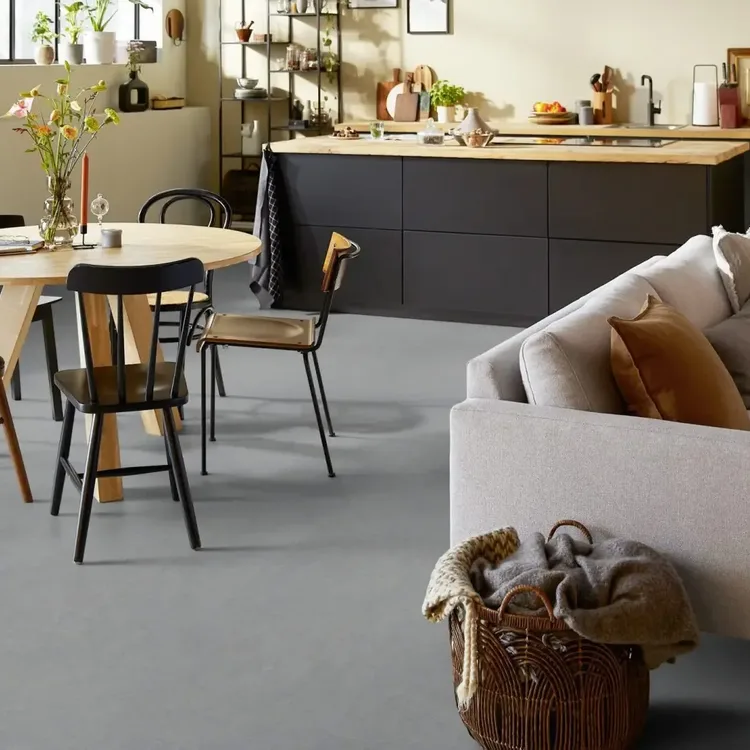 Open woonkeuken met grijze marmoleumvloer, ronde houten eettafel, lichte bank en zwart keukeneiland
